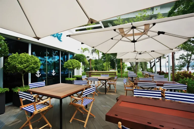 Outdoor café with striped chairs, wooden tables, and large white umbrellas. Surrounded by greenery, the setting feels inviting and serene.