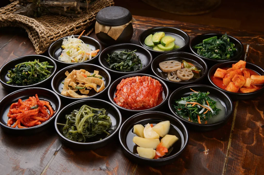 A variety of colorful Korean side dishes, or banchan, in small black bowls on a wooden table. Kimchi, pickles, and vegetables showcased, creating a vibrant spread.