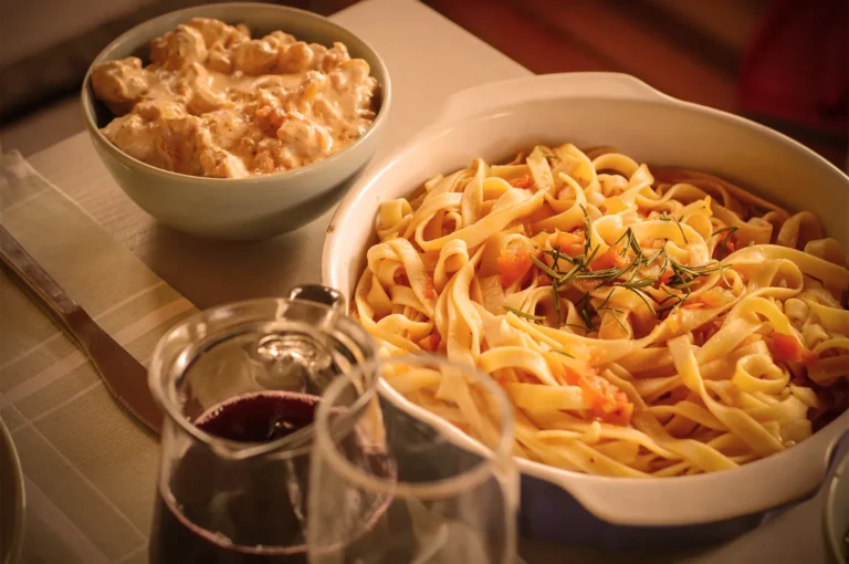 A cozy dinner scene featuring a bowl of creamy pasta topped with herbs, a side dish of dumplings, and a glass of red wine, set on a softly lit table.