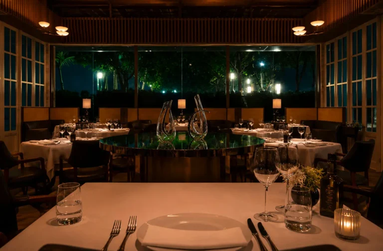 Elegant fine dining restaurant interior at night with white tablecloths, wine glasses, and garden views through floor-to-ceiling windows