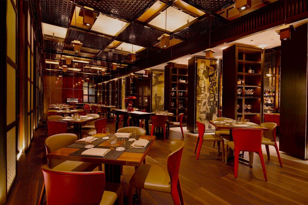 Elegant restaurant interior with warm lighting, red and beige chairs, wooden tables set for dining. A cozy, modern ambiance with Asian-inspired decor.