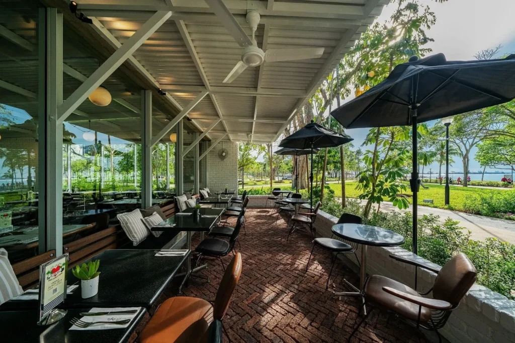 Outdoor café seating with black tables, cushioned chairs, and large umbrellas. Brick flooring, lush greenery, and waterfront view create a serene ambiance.