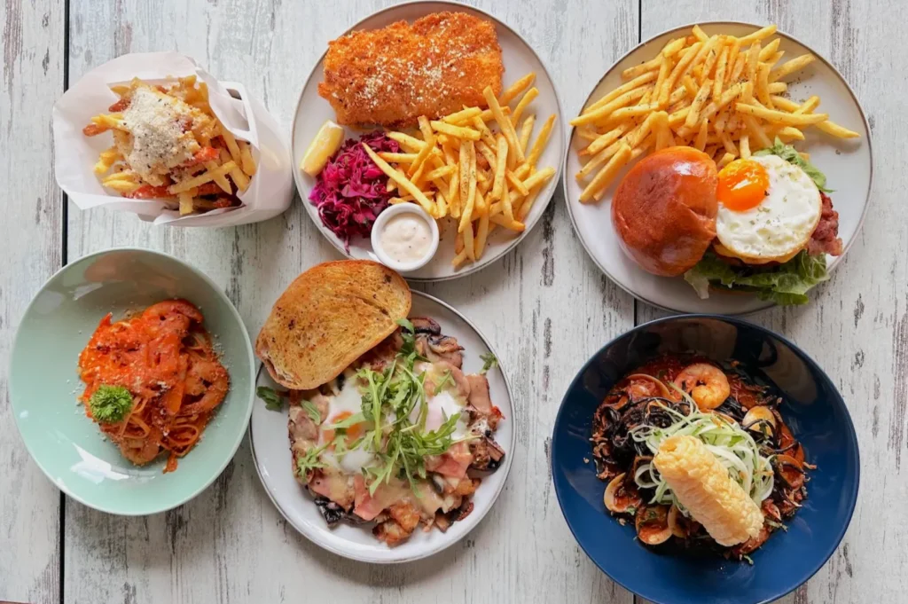 Aerial view of a wooden table with diverse dishes: fries, schnitzel with purple slaw, burger with egg, seafood pasta, and more, creating a vibrant, appetizing spread.