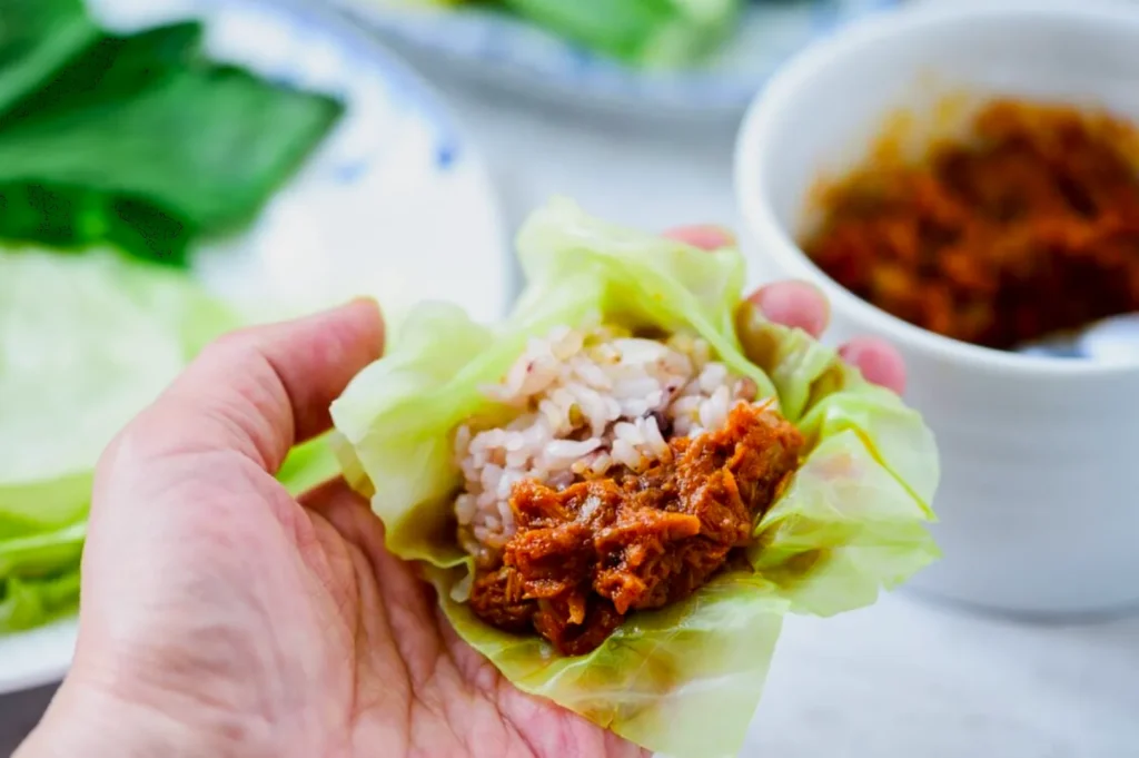 A hand holds a cabbage leaf filled with rice and a reddish-brown meat sauce. In the background, there's a bowl and more cabbage leaves, creating a fresh and appetizing tone.