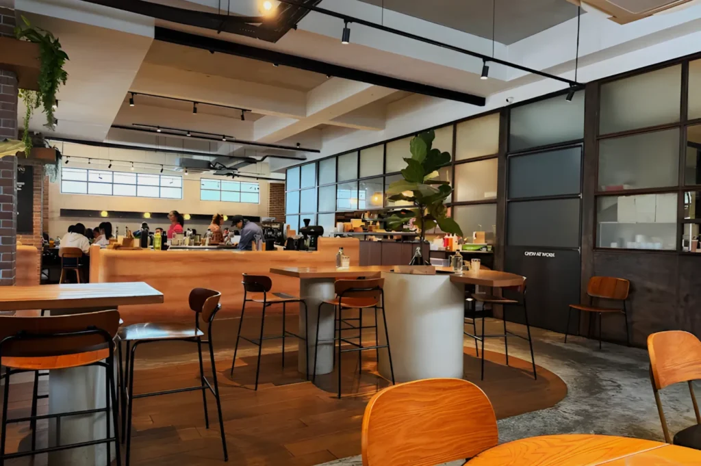 Modern cafe interior with wooden furniture, high tables, and a cozy atmosphere. Large frosted windows and plants add warmth. Patrons enjoy drinks.