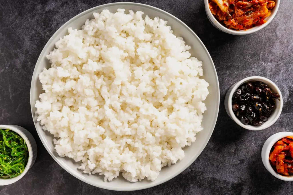 A bowl of fluffy white rice is centered, surrounded by small bowls of seaweed, pickles, and kimchi on a dark textured surface, creating a savory and inviting scene.