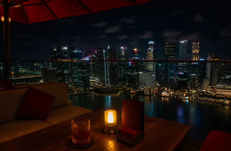 Rooftop bar at night with cocktail and candle on table overlooking illuminated Singapore skyline and Marina Bay waterfront.