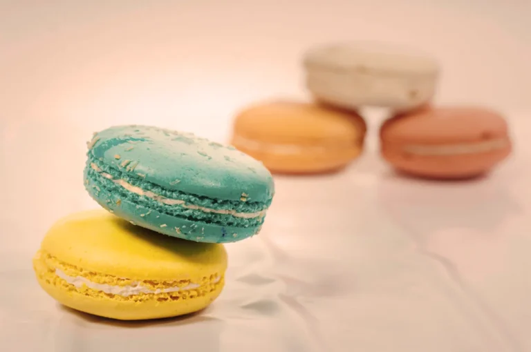 Stacked colorful macarons are pictured with blue and yellow ones in focus, and out-of-focus peach, pink, and white ones in the background.