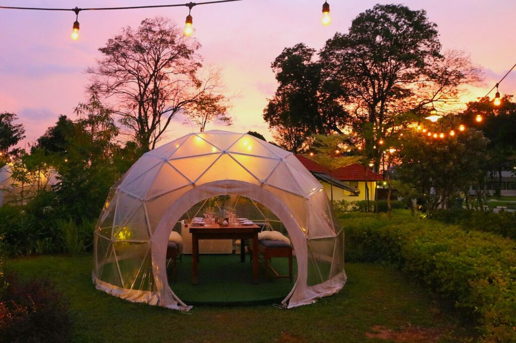 A clear geodesic dome sits on a grassy area at sunset, with a table set for dining inside. Warm string lights hang above, creating a cozy ambiance.