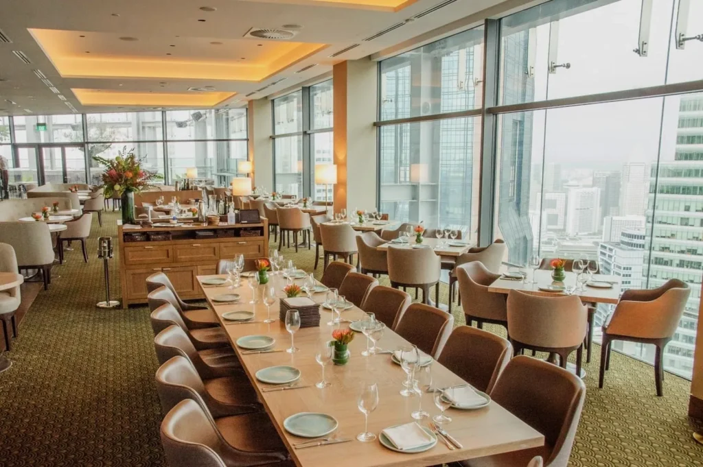 Elegant rooftop restaurant with large windows offering a city view. Tables are set with white plates, glasses, and orange flower centerpieces.
