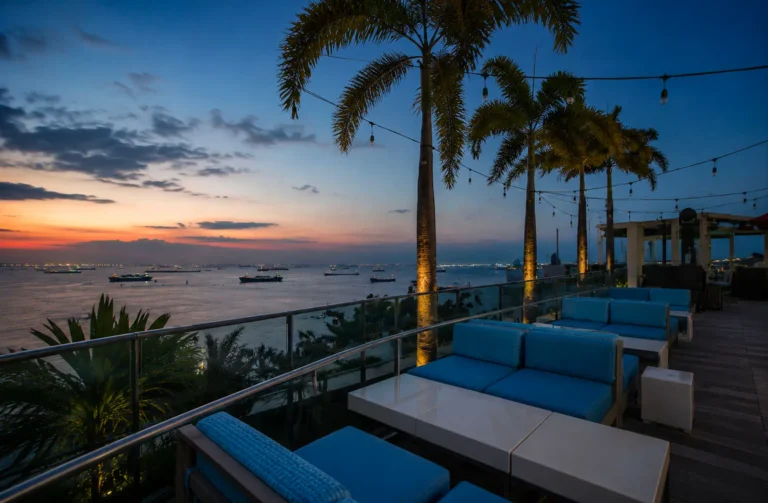 Rooftop lounge with blue cushioned seating and palm trees overlooking harbor at sunset with cargo ships and twilight sky