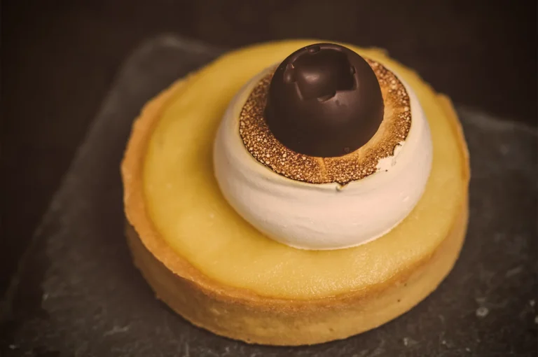 A lemon tart with a smooth, yellow filling is topped with toasted meringue and a dark chocolate sphere, creating an elegant, gourmet dessert look.