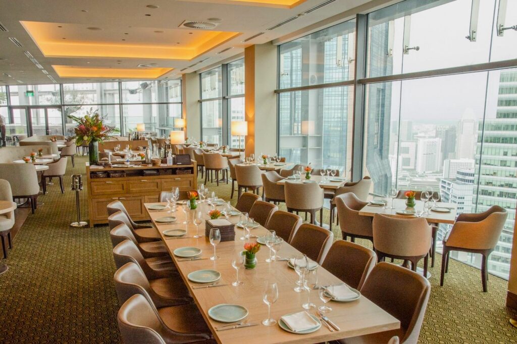 Upscale restaurant with cityscape views. Elegant tables set with wine glasses, white plates, and small flower vases. Large windows and warm lighting.