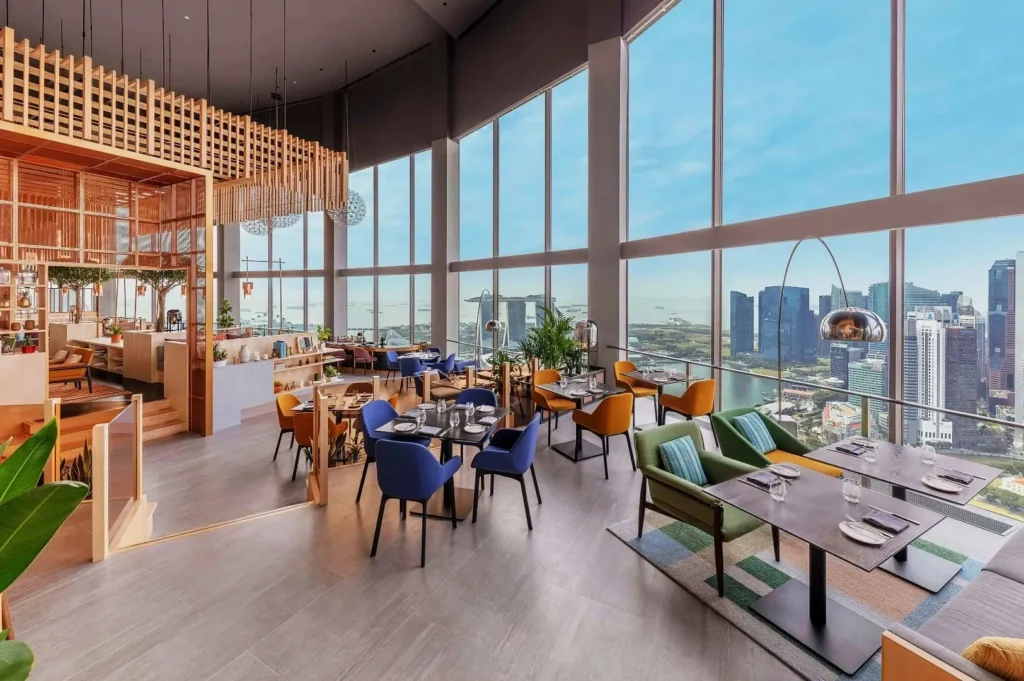 Spacious, modern restaurant with tall windows showcasing a city skyline. Features colorful chairs, wooden decor, and a vibrant, airy ambiance.