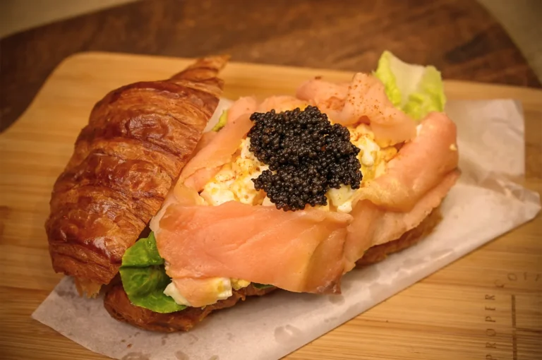 A croissant sandwich on a wooden board, filled with lettuce, scrambled eggs, smoked salmon, and topped with a spoonful of black caviar. The image conveys luxury.