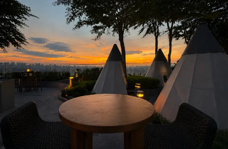 Rooftop terrace with teepee-style tent structures and wooden table overlooking city skyline at sunset with orange and blue sky