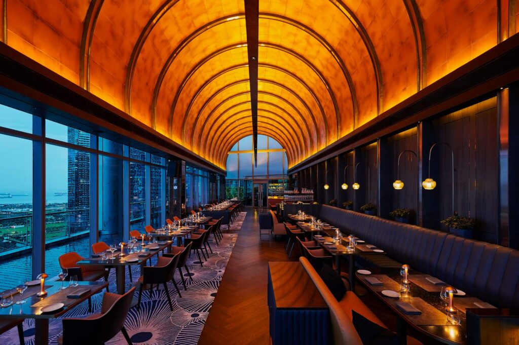 Elegant restaurant interior with arched, glowing ceiling. Floor-to-ceiling windows reveal city and waterfront views. Tables are set for dining.