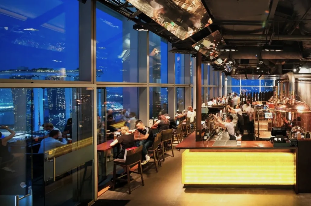 A lively bar scene at night with people seated at tables by large windows overlooking a cityscape. Warm lighting and polished interiors create an inviting atmosphere.