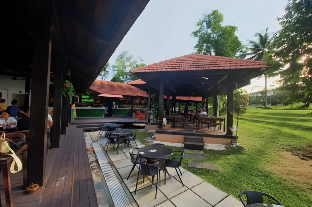 Outdoor dining area with open-air gazebos featuring red tiled roofs, wooden decks, and round tables. Lush green grass and trees create a serene atmosphere.