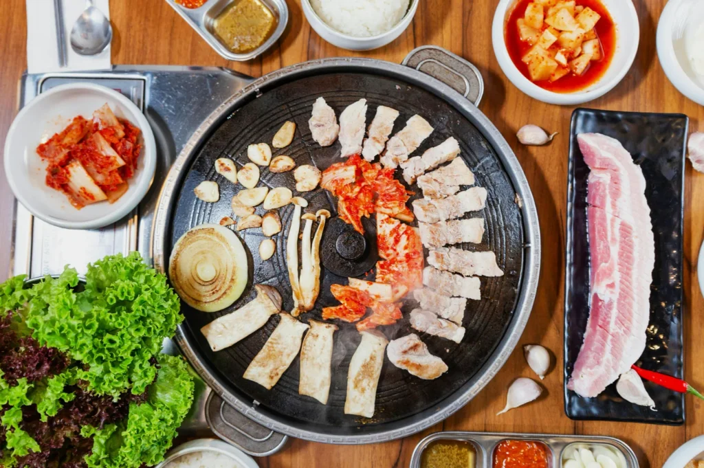 A Korean barbecue grill loaded with sizzling meat, kimchi, garlic, mushrooms, and onions. Surrounding dishes include fresh lettuce, condiments, and kimchi.