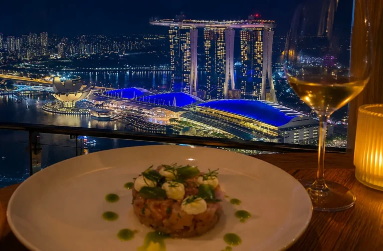 Gourmet tuna tartare dish with white wine on rooftop dining table overlooking illuminated Marina Bay Sands and Singapore skyline at night