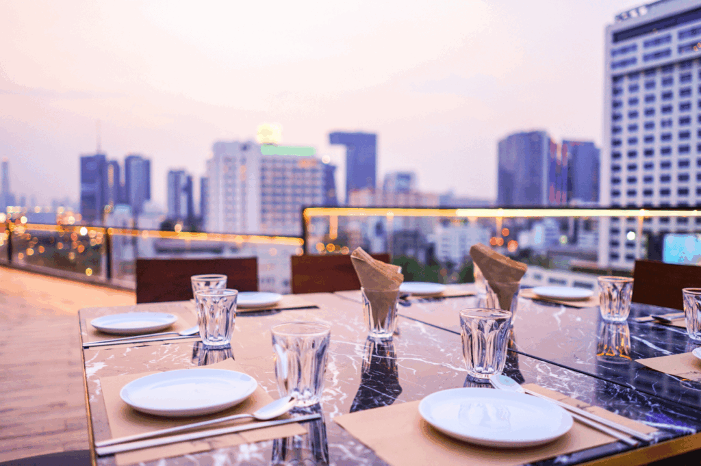 Elegant rooftop dining setup overlooking Singapore’s cityscape at dusk, featuring a marble table set for a private gathering with folded brown napkins, white plates, and crystal-clear glassware—evoking intimacy, modern luxury, and the allure of urban hospitality.