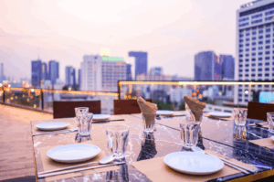 Elegant rooftop dining setup overlooking Singapore’s cityscape at dusk, featuring a marble table set for a private gathering with folded brown napkins, white plates, and crystal-clear glassware—evoking intimacy, modern luxury, and the allure of urban hospitality.