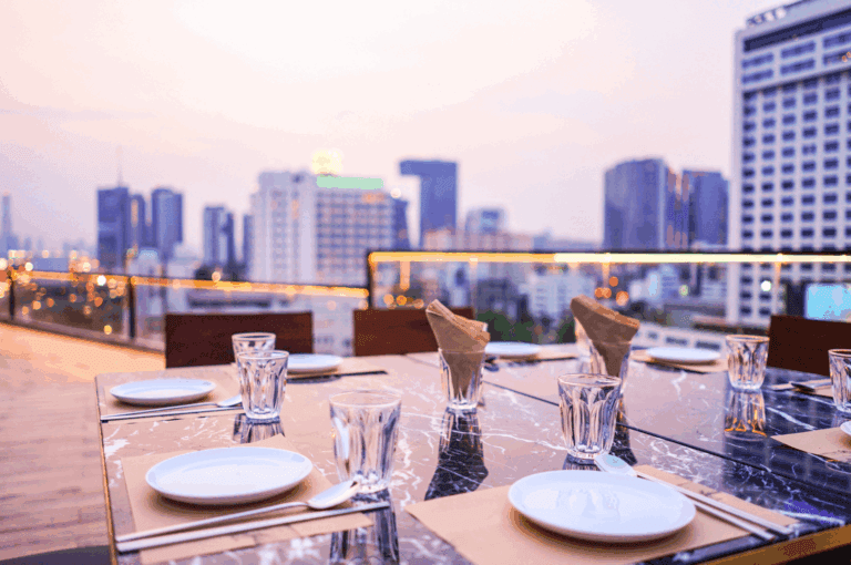 Elegant rooftop dining setup overlooking Singapore’s cityscape at dusk, featuring a marble table set for a private gathering with folded brown napkins, white plates, and crystal-clear glassware—evoking intimacy, modern luxury, and the allure of urban hospitality.