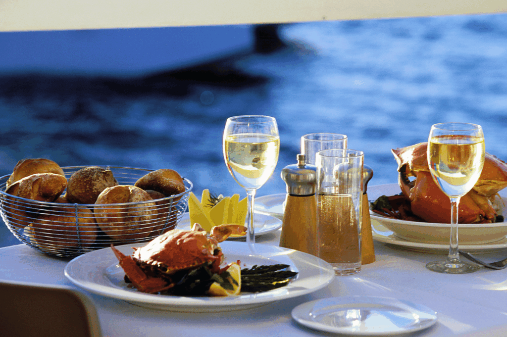 Waterfront fine dining table set for two, featuring crab dishes with lemon wedges, fresh bread rolls, and white wine—capturing the serenity and indulgence of a private culinary escape by the sea.
