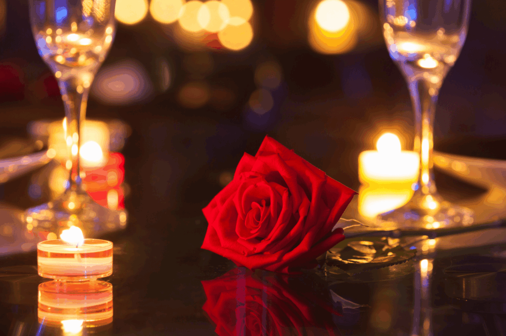Intimate celebration scene with a red rose, flickering tealight candles, and empty champagne flutes on a reflective surface—suggesting romance, anticipation, and the quiet elegance of a private toast.