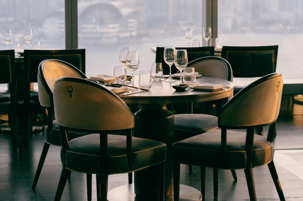Stylish round dining table set for four with wine glasses, plush chairs, and a panoramic city-and-waterfront view through floor-to-ceiling windows—evoking serene sophistication and the exclusivity of high-rise private dining.