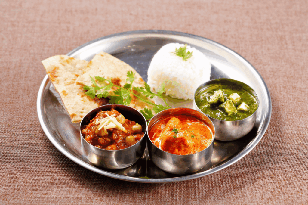 Traditional Indian thali served on a stainless steel plate with white rice, cilantro-garnished naan, and three vibrant curries—chickpea masala, creamy tomato-based paneer, and spinach-paneer palak—capturing the authentic flavors and balanced textures celebrated in Little India, Singapore.