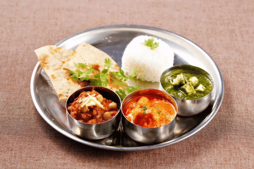 Traditional Indian thali served on a stainless steel plate with white rice, cilantro-garnished naan, and three vibrant curries—chickpea masala, creamy tomato-based paneer, and spinach-paneer palak—capturing the authentic flavors and balanced textures celebrated in Little India, Singapore.