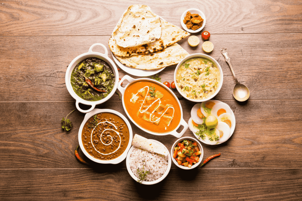 Elegant Indian meal spread on a wooden surface featuring palak paneer, dal, butter chicken, khichdi, white rice with papadum, assorted sweets, salad, naan, and condiments—showcasing the comforting depth and festive variety of Little India’s culinary traditions in Singapore.