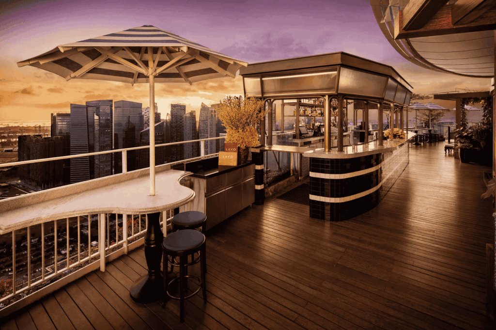 Elegant romantic rooftop dining Singapore setting at a rooftop bar with wooden deck, bar stools, tables, and umbrellas overlooking the Singapore skyline at sunset, offering stunning views of Marina Bay, Marina Bay Sands, and the Singapore Flyer. Perfect for a romantic date night, corporate events, or celebrations, this restaurant in Singapore ranks among the most romantic restaurants and rooftop restaurants, serving fine dining experiences that blend contemporary Asian cuisine with Italian restaurant classics. Guests enjoy great food, steaks, wine, signature cocktails, and an imaginative menu by an executive chef, paired with impeccable service and a la carte dishes from a new menu. The city skyline and city lights create breathtaking views and skyline views at sea level, with the location at Collyer Quay surrounded by world‑class restaurants, bar spaces, and dining options. Whether for lunch, dinner, afternoon bites, or evening occasions, friends and couples sip drinks, explore flavours, and feast on quality food in an elegant yet casual space. Every meal is a blend of taste, fun, and delight, with each bite a note of flavour against a backdrop that invites you to soak in the moment, miss nothing, and literally explore the best dishes, cocktails, and events Singapore has to offer, with access to the east and tickets to unforgettable occasions.