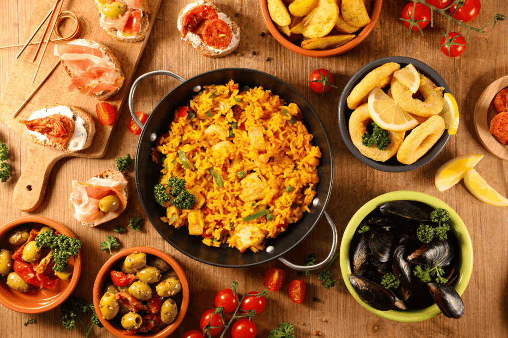 A colorful feast of Spanish cuisine and Spanish food featuring traditional Spanish dishes and every beloved Spanish dish from seafood paella—the national dish and most famous Spanish dish—to tapas like cured ham, chorizo, black pudding, blood sausage, and pimientos de padrón (de padrón). This rustic table showcases traditional Spanish recipes and Spanish classics such as Spanish omelette, patatas bravas with tomato sauce, fresh bread, crusty bread drizzled with olive oil, and popular tapa plates served hot for a dinner party with loved ones. Surrounding the paella are rice dishes, meat and chicken options, potatoes, green peppers, deep fried calamari, mussels, and other ingredients like garlic and mojo picón from the Canary Islands and Canarian cuisine. Regional flavors from Basque Country, La Rioja, and the Iberian Peninsula appear alongside rabo de toro (braised oxtail), pork fat-enriched stews, ground beef dishes, and cold soup. Perfect for those who visit Spain, dine at a restaurant like El Celler de Can Roca (El Celler), or are a huge fan of Spanish cooking. The delicious meal ends with popular desserts and dessert treats such as chocolate creations and dairy products, offering a rich flavor experience that invites you to eat, taste, and celebrate food, cuisine, and traditional dishes from Spain.