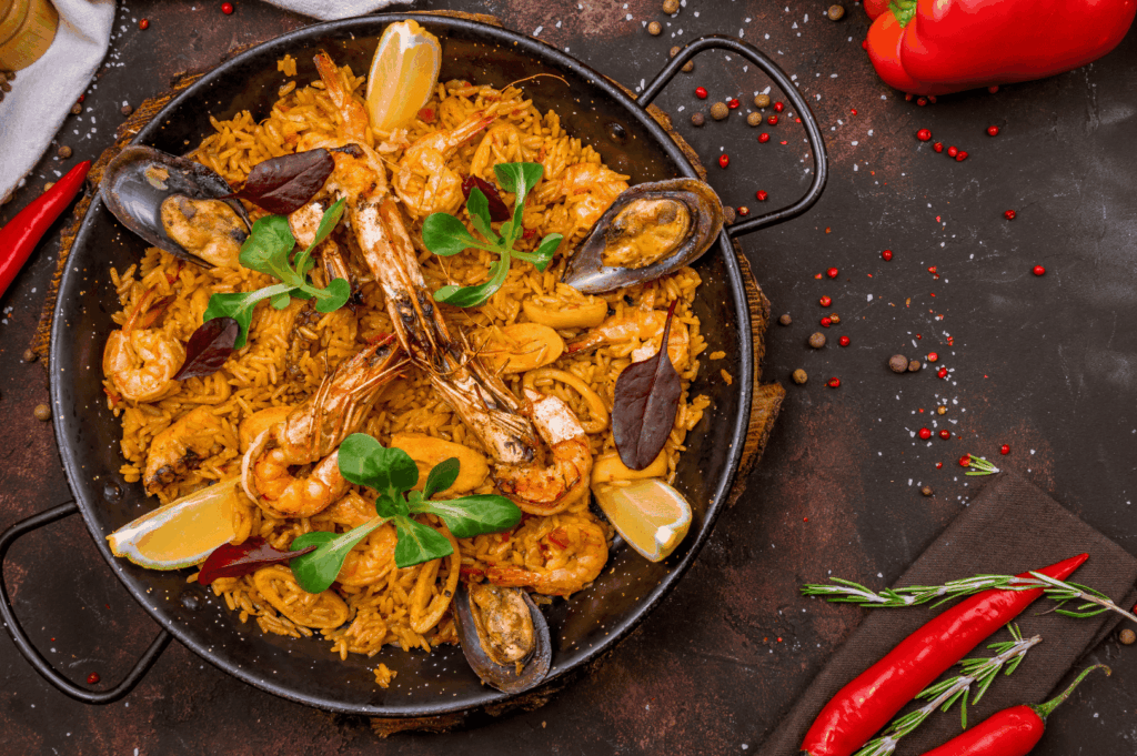 A vibrant seafood paella — the national dish of Spain and one of the most famous Spanish dishes — served hot in a black pan, brimming with golden rice cooked in olive oil, plump shrimp, mussels, green peppers, and garlic, garnished with lemon wedges and fresh herbs. This famous Spanish dish, a jewel of Spanish cuisine and Spanish food, represents traditional Spanish dishes and traditional Spanish recipes from the Iberian Peninsula, beloved in Spanish cooking and celebrated as the ultimate Spanish comfort food. Around the pan, fresh bread and crusty bread evoke popular tapa culture, alongside ingredients like tomato sauce, chorizo, meat, chicken, pork fat, ground beef, blood sausage, black pudding, and cured ham — staples of traditional dishes and Spanish classics such as Spanish omelette, patatas bravas, rabo de toro, braised oxtail, pimientos de padrón, and mojo picón from the Canary Islands and Canarian cuisine. This dish reflects flavors from Basque Country, La Rioja, and El Celler de traditions, perfect for a dinner party with loved ones or to eat at a restaurant when you visit Spain. It shares the table with popular desserts like chocolate and dessert pastries, deep fried treats, and dairy products, showing the diversity of cuisine from white rice rice dishes to potatoes, tapas, and other ingredients that define traditional Spanish and popular dishes. A feast of rich flavor, delicious textures, and authentic taste, this food celebrates the artistry of Spanish gastronomy and the joy of a shared meal.