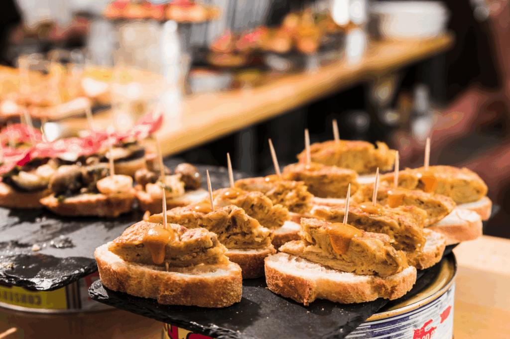 Close‑up of a popular tapa from Spanish cuisine, showcasing fresh bread and crusty bread topped with slices of chorizo, blood sausage, or black pudding, drizzled with tomato sauce and olive oil, served hot on a rustic slate tray for a dinner party in Spain. This Spanish dish, part of traditional Spanish dishes and traditional dishes rooted in traditional Spanish recipes, reflects Spanish cooking from the Basque Country to the Canary Islands and La Rioja. A famous Spanish dish beloved by a huge fan of Spanish food, it pairs well with seafood paella, paella, rice dishes, white rice, potatoes, patatas bravas, spanish omelette, pimientos de padrón, de padrón, mojo picón, garlic, and other ingredients from canarian cuisine. Inspired by el celler and el celler de traditions, it embodies spanish classics like rabo de toro, braised oxtail, cured ham, ground beef, green peppers, pork fat, meat, chicken, seafood, and deep fried specialties from the iberian peninsula. Perfect to eat and taste with loved ones at a restaurant when you visit spain, this national dish offers rich flavor and the ultimate Spanish comfort food experience. Complements popular dishes, popular desserts, dessert, chocolate, dairy products, and meal traditions across spain.