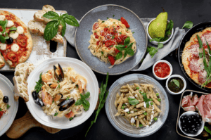 A beautifully arranged spread of Italian cuisine and Italian food featuring authentic Italian cuisine classics such as wood-fired pizza topped with mozzarella cheese, prosciutto, ripe tomatoes, oregano, and fresh vegetables, alongside Italian dishes of fresh pasta and pasta varieties like fettuccine with tomato sauce, meat sauce, pesto sauce, and parmesan cheese. The traditional dish selection includes stuffed pasta, seafood pasta with lemon juice, butter, and herbs, and baked main courses made from wheat flour, buffalo milk, and extra virgin olive oil. Garnished with pine nuts, garlic, basil, and balsamic vinegar, these iconic dishes reflect the long history of Italy’s northern Italy, southern Italy, and Emilia Romagna regions, where flavors vary depending on the primary ingredient. Served with rustic bread, potatoes, beef, chicken, cannellini beans, and vegetables, plus indulgent desserts and desserts typically served in an Italian restaurant. Styled with expert food styling to highlight great food, various sauces, and many variations of dishes, this meal showcases a deep connection to recipes and cooked traditions, celebrating cheese, milk, salt, mozzarella, sauce, meat, dough, and risotto as the heart of Italian food culture.