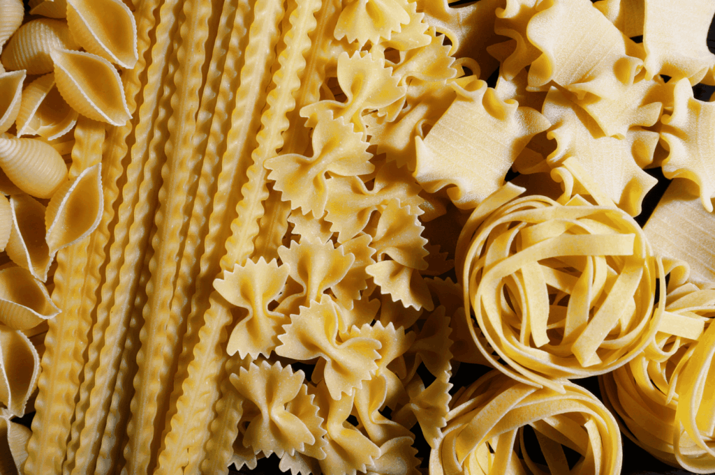 A beautifully arranged selection of uncooked pasta varieties representing the heart of Italian cuisine and Italian food, including conchiglie, farfalle, mafaldine, lasagnette, and tagliatelle nests made from wheat flour, dough, and primary ingredients like buffalo milk, eggs, and salt. These fresh pasta shapes, typically served in Italian dishes at an Italian restaurant, are the foundation for traditional dish recipes from northern Italy, southern Italy, and Emilia Romagna, where flavors vary depending on the sauce and main courses. Perfect for pairing with tomato sauce, meat sauce, pesto sauce, parmesan cheese, mozzarella cheese, mozzarella, prosciutto, seafood, chicken, beef, vegetables, fresh vegetables, ripe tomatoes, tomatoes, garlic, herbs, oregano, pine nuts, olive oil, and extra virgin olive oil. Complements iconic dishes like stuffed pasta, risotto, pizza, baked meals, and indulgent desserts or desserts served with bread, potatoes, cannellini beans, and great food from Italy. Styled with expert food styling to highlight many variations, various sauces, and the deep connection to recipes, cooked traditions, and the long history of Italian culinary culture, enhanced with touches of balsamic vinegar, lemon juice, lemon, butter, melted butter, milk, and cheese for a complete meal.