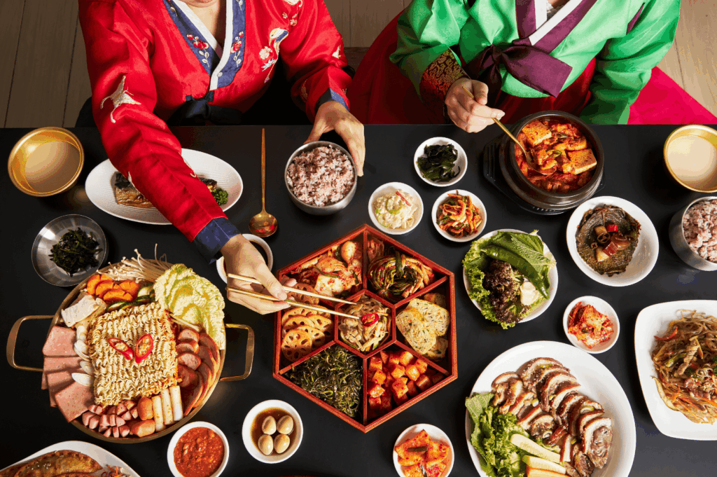 Traditional Korean meal spread featuring comforting Korean dishes like kimchi stew, grilled meats, multigrain rice, and assorted side dishes served in a hexagonal tray, enjoyed by diners in colorful hanbok attire. This vibrant Korean cuisine experience, ideal for special occasions and sharing platters, reflects the authentic flavors found at top Korean restaurants in Singapore such as Charim Korean BBQ, Wang Dae Bak, and Hanjip Korean Grill House. Perfect for la carte menu options, set menus, and private dining rooms, the spread includes pork belly, beef brisket, steamed dumplings, and rice cakes—highlighting the best Korean BBQ and Mul Gogi Korean BBQ restaurant offerings with premium meat cuts and attentive service.