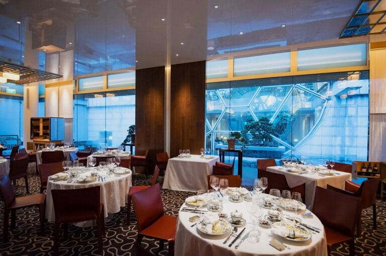 Upscale restaurant interior; round tables with white tablecloths, elegant place settings, and wine glasses. Large windows reveal a geometric outdoor view.