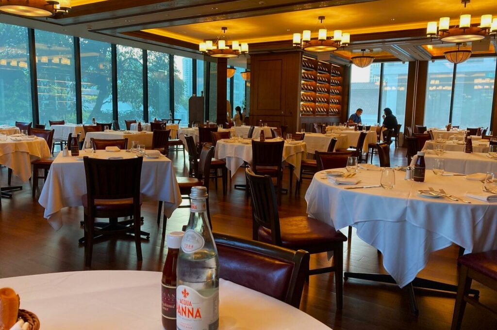 Elegant restaurant interior with round tables covered in white tablecloths, set with glassware. Large windows offer natural light, creating a cozy ambiance.