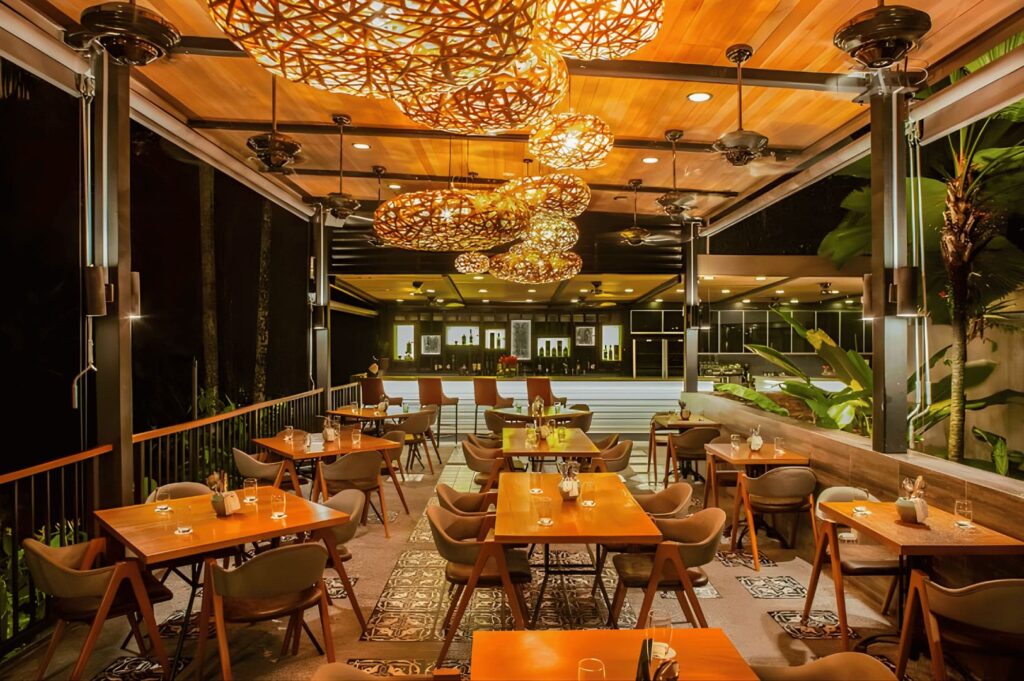 Warmly lit outdoor restaurant with wooden tables and modern chairs, featuring intricate hanging lights. The ambiance is cozy and inviting.