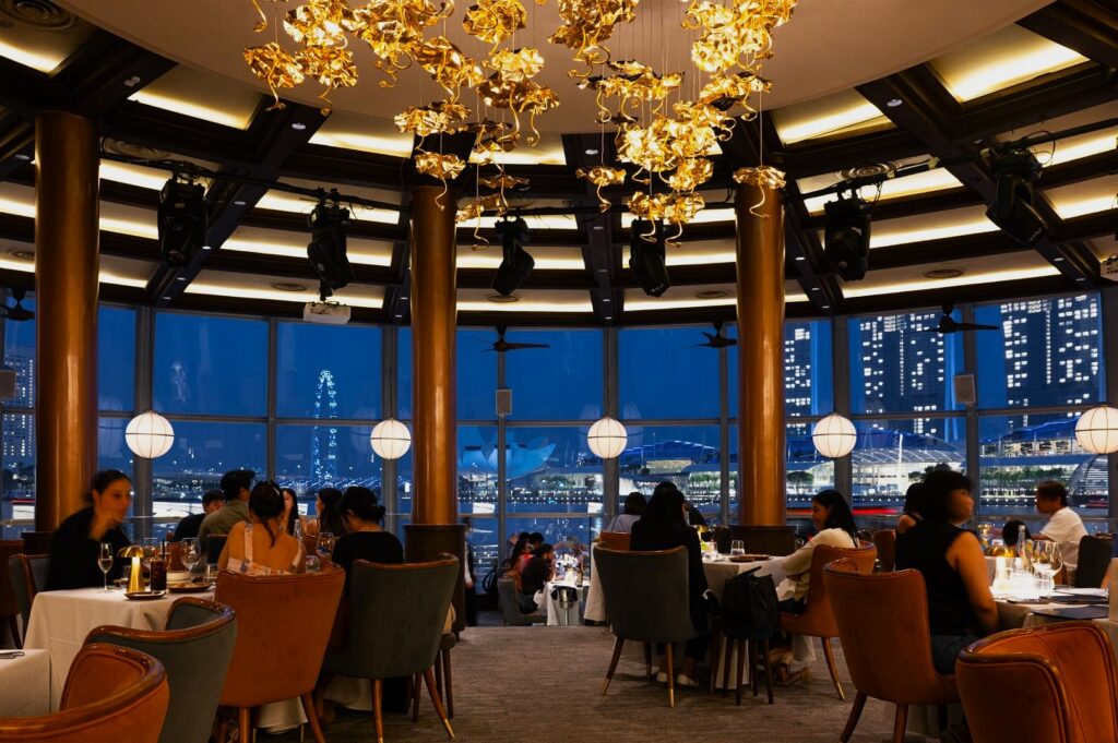 Elegant restaurant with modern decor, featuring golden chandeliers and floor-to-ceiling windows. Patrons dine at round tables, with a night cityscape visible.