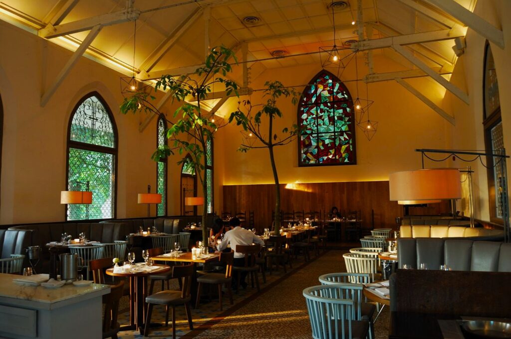 Spacious restaurant with high ceilings, stained glass window, and cozy lighting. Diners sit at wooden tables and chairs, creating an elegant, warm atmosphere.