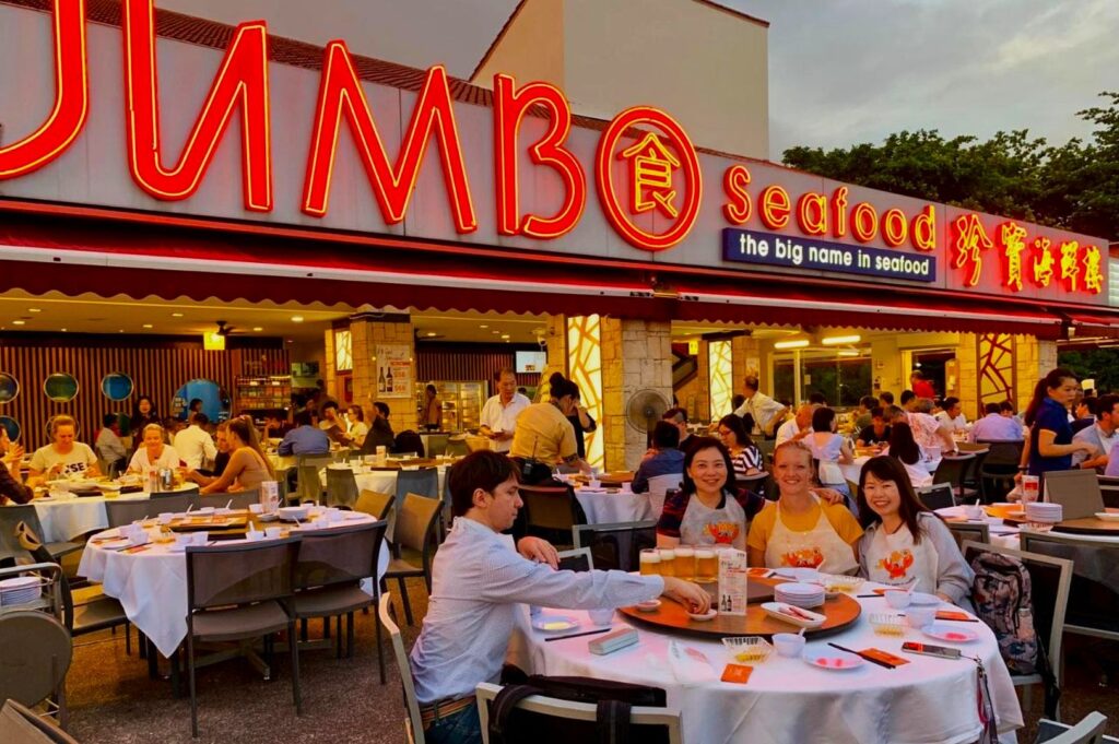 Outdoor dining at Jumbo Seafood, with neon signage in the background. People are enjoying a meal at round tables, exuding a lively, social atmosphere.