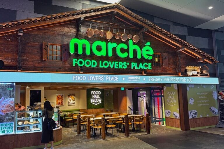 Rustic-themed restaurant exterior with wooden decor and green neon sign reading "Marché Food Lovers' Place." A person stands by a dessert counter.