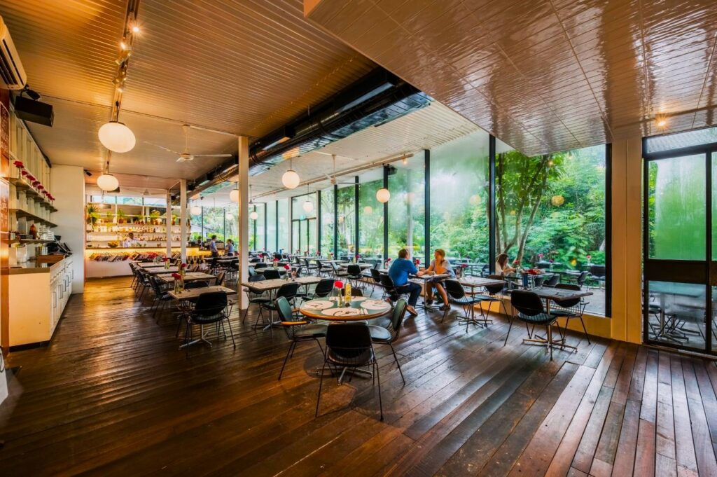Spacious restaurant interior with wooden floors, modern round tables, large windows displaying lush greenery outside, and a few diners seated.