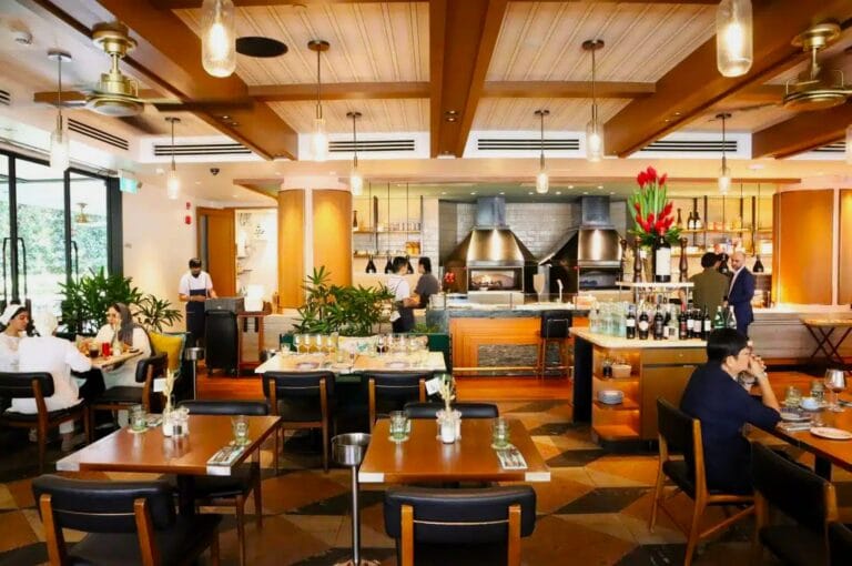 Modern restaurant interior with wooden beams, pendant lighting, and green plants. Diners sit at wooden tables, while chefs work in an open kitchen. Cozy and inviting atmosphere.